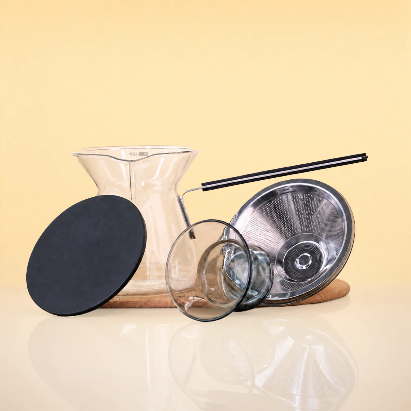 ObsidianEditionCeremonySet2 - A glass pour-over coffee maker set with a clear glass carafe, glass filter cone, glass cup, black metal strainer, black handle, and a round black lid, arranged on a wooden coaster against a warm yellow background.