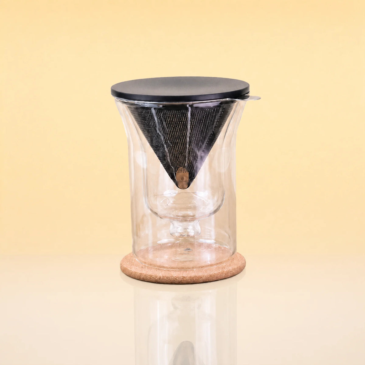 TeaChalice-DoubleWalledGlass_TeaInfuserSet - A clear glass pour-over coffee dripper with a black mesh cone filter, a black lid, and a round cork base, shown on a warm beige background.