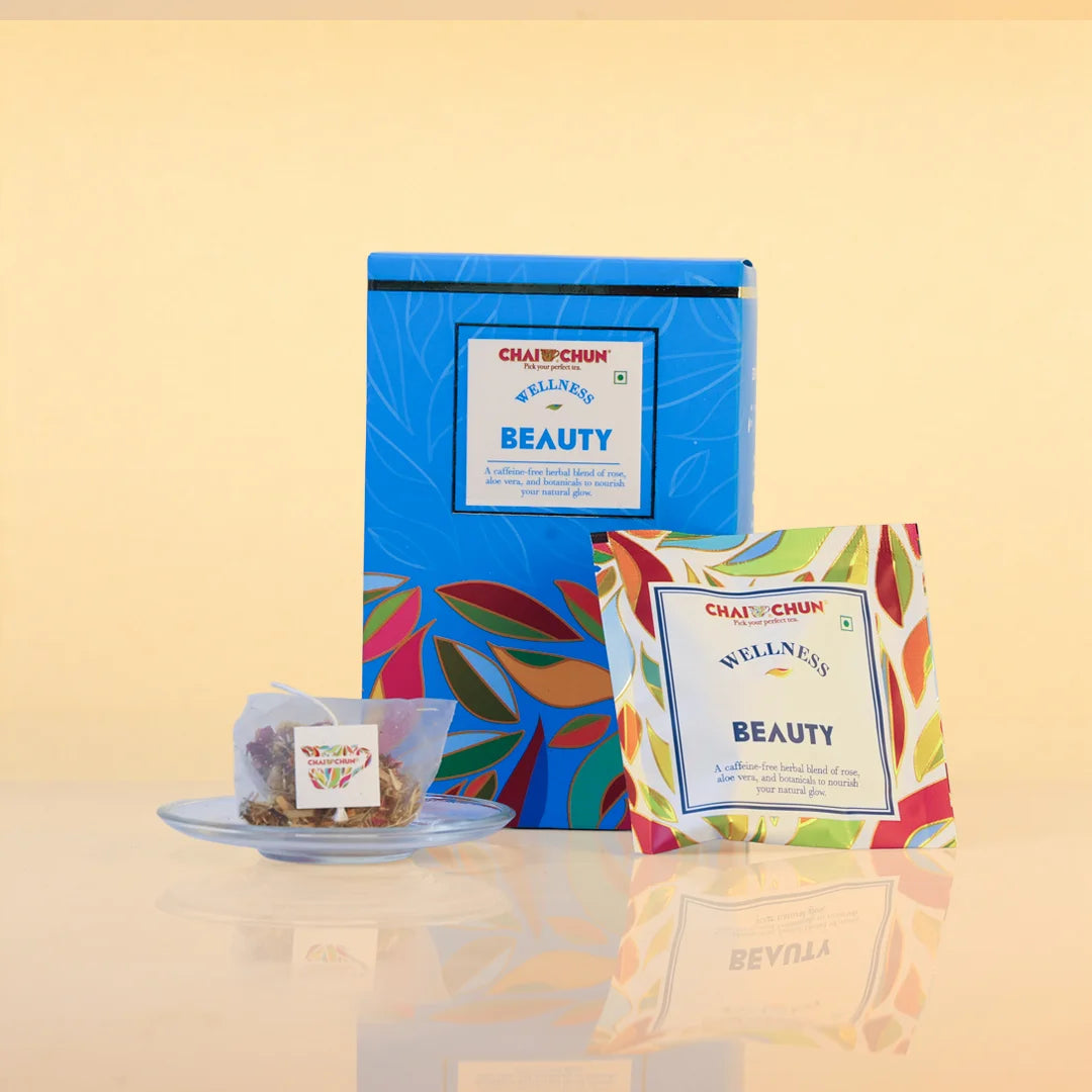 Chai Chai Chan 'Wellness with Beauty' tea set featuring a blue box, a single tea bag on a small saucer, and a single sachet with colorful leaf patterns against a warm beige background.