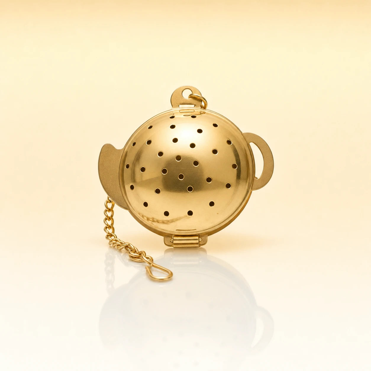 Turtle Bloom Infuser – Gold-toned stainless steel ball-shaped tea infuser with small perforations, side handles, and a gold chain with clasp, photographed on a warm cream background with a soft reflection below.