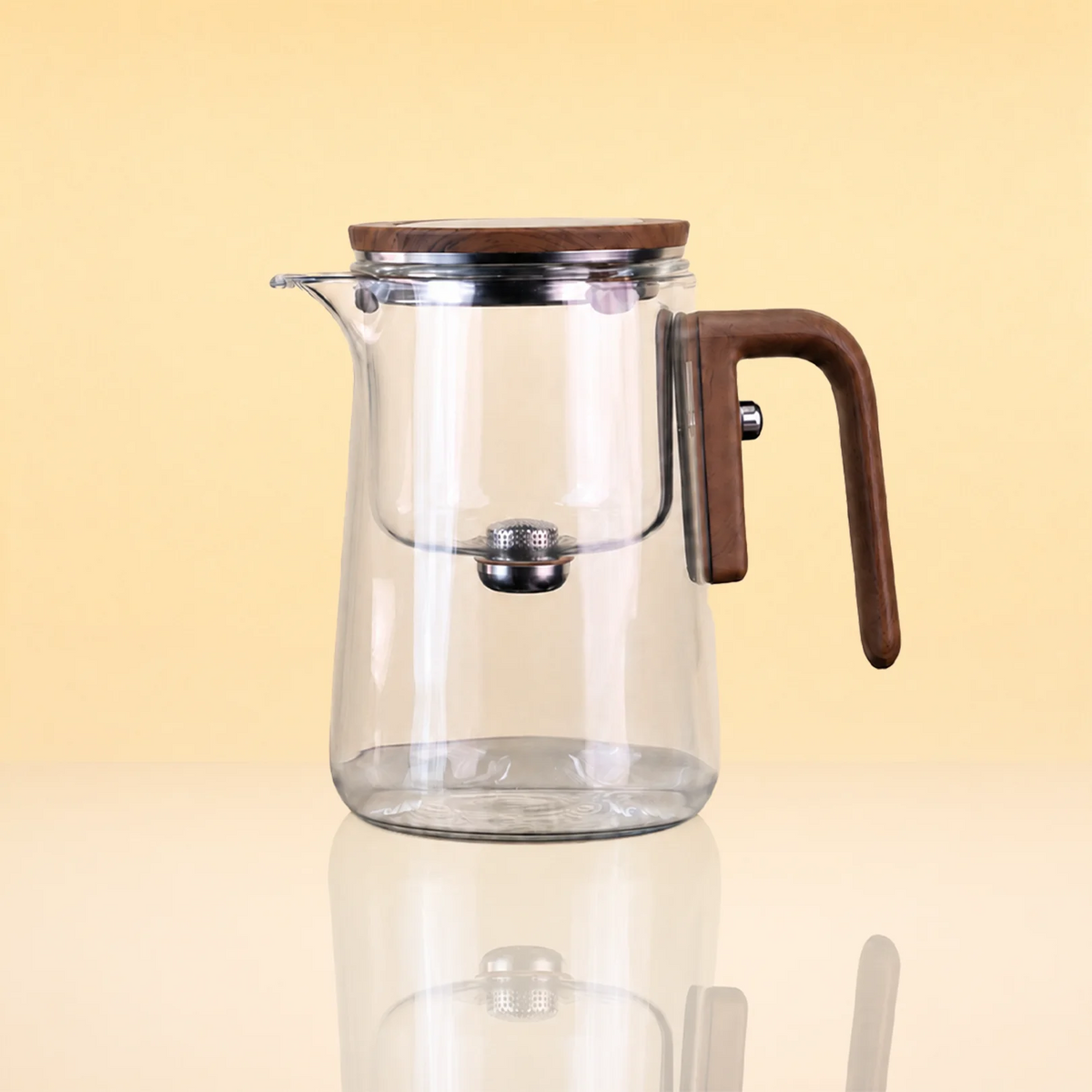 AlchemyMagneticGlassTeapot - Clear glass teapot with a brown wooden lid and handle, and a stainless steel infuser, shown on a reflective beige surface against a warm yellow background.