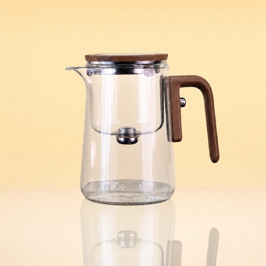 AlchemyMagneticGlassTeapot - Clear glass teapot with a brown wooden lid and handle, and a stainless steel infuser, shown on a reflective beige surface against a warm yellow background.