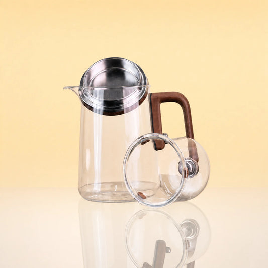 Alchemy_Magnetic_Glass_Teapot_2 - Clear glass teapot with a stainless steel lid and a brown wooden handle, shown with a matching glass cup on a warm beige background.