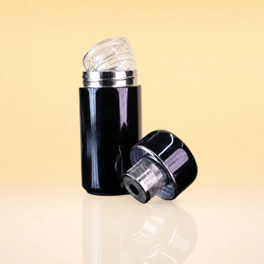 AuraFlask-TeaInfusingTumbler_small - A glossy black cylindrical bottle or container with a clear ribbed cap, shown with the cap removed and leaning beside it on a warm beige studio background.