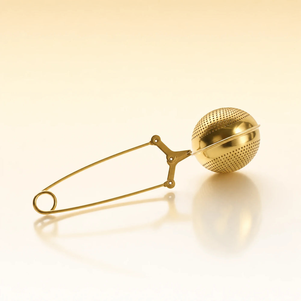 Aura Ball Infuser – Gold-toned stainless steel spherical tea infuser with a textured perforated surface, attached to a long safety-pin style clasp, photographed on a warm cream background with a soft reflection below.