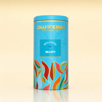 A cylindrical blue canister of Chai Chuk Wellness Yoga Tea, featuring colorful leaf illustrations and gold lettering, set against a warm beige background with a soft reflection below.