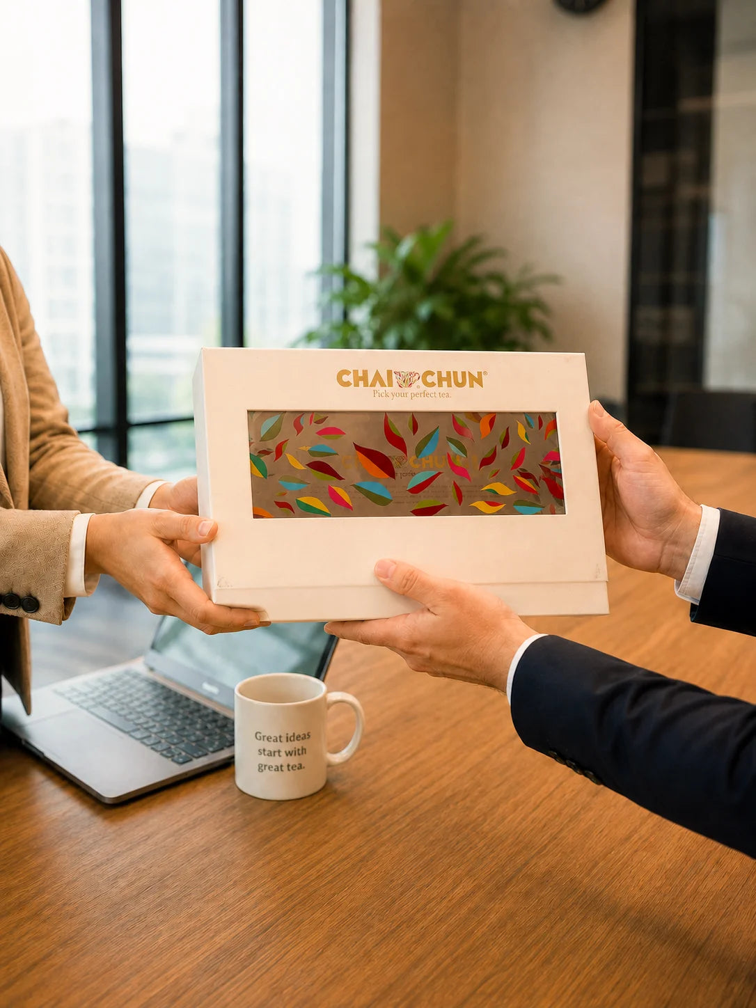 CORPORATE-GIFTING - A person in a beige blazer and another in a dark suit hold a premium white box labeled CHAICHUN Tea over a wooden table in a modern office setting. A white mug with text sits on the table, along with a laptop and a blurred plant in the background.