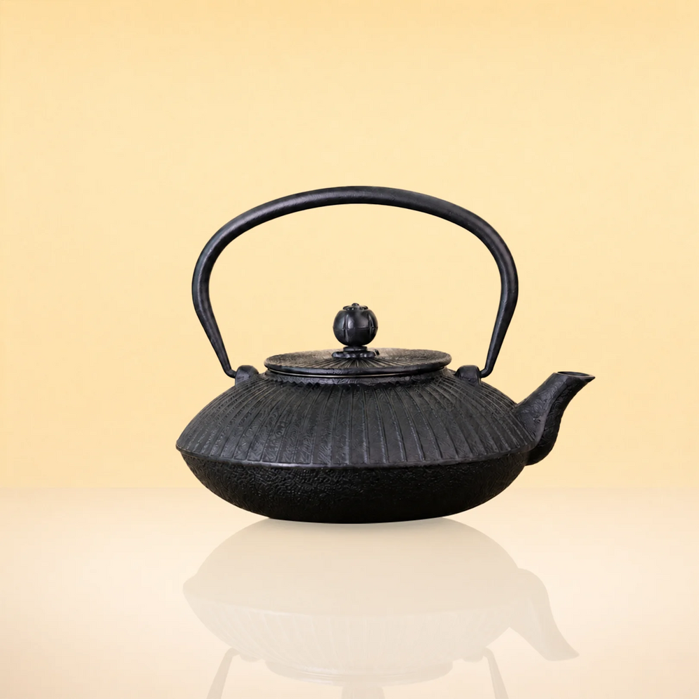 CharcoalInfusionFlutedIronTeapot A black cast iron teapot with a ribbed textured body, curved handle, and small spout, centered on a warm beige gradient background with a soft reflection below.