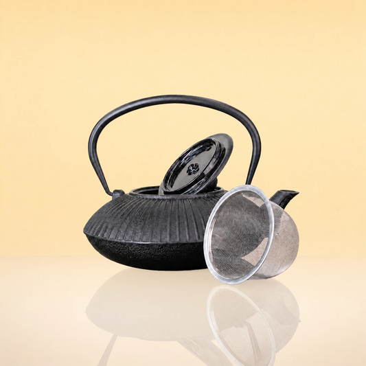 CharcoalInfusionFlutedIronTeapot2 - A black cast iron teapot with a ribbed textured body, a curved handle, a matching black lid, and a clear mesh strainer resting beside it on a reflective surface against a warm beige background.