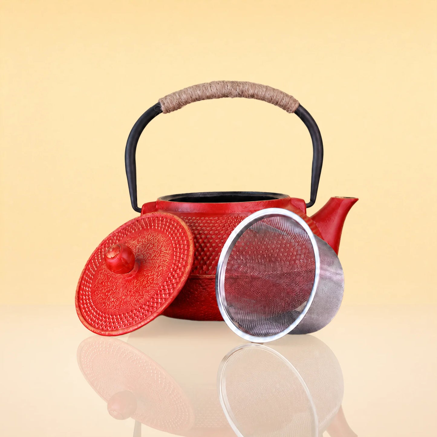 CrimsonBloomIronTeapot2 - A red textured cast-iron teapot with a black handle and a beige wrapped grip, shown with its matching red lid and a clear mesh strainer on a warm beige reflective surface.