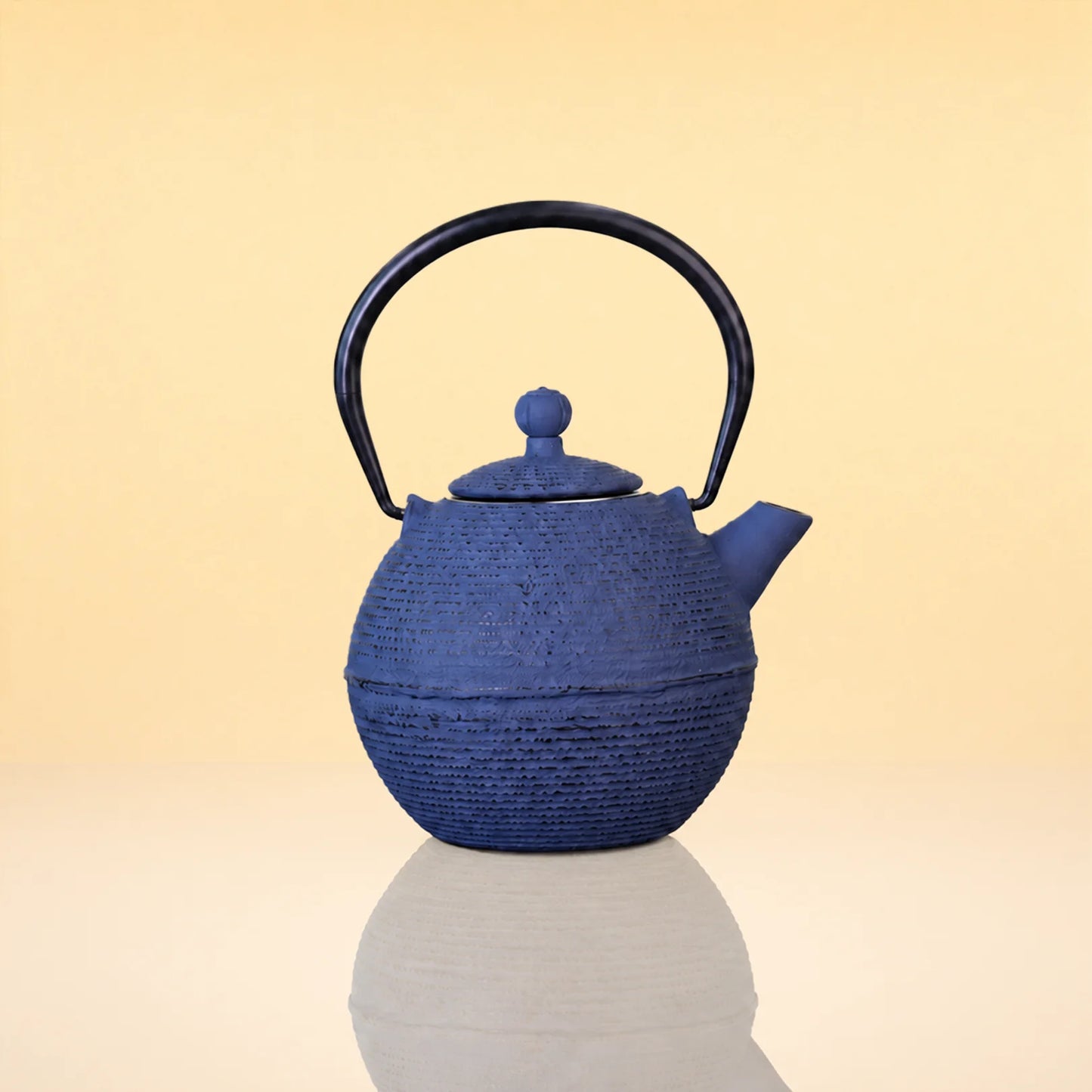 DuskPebbleIronTeapot - A textured blue cast iron teapot with a black metal handle, shown on a reflective surface against a warm beige gradient background.