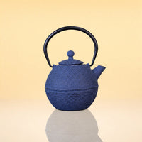 DuskPebbleIronTeapot - A textured blue cast iron teapot with a black metal handle, shown on a reflective surface against a warm beige gradient background.
