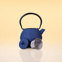 DuskPebbleIronTeapot2 - A textured dark blue cast iron teapot with a black handle, shown with its lid off and a metal mesh strainer on a reflective beige surface.
