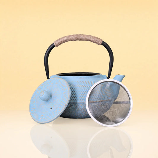 FrostArcIronTeapot - A light blue textured cast iron teapot with a black handle wrapped in beige cord, shown with its lid off and a metal tea strainer on a reflective cream surface against a pale yellow background.