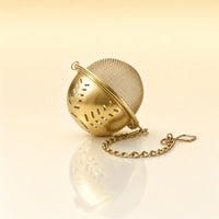 GOLDEN LEAF STRAINER - A gold-finished metal tea infuser ball with a matching chain, resting on a reflective cream surface.