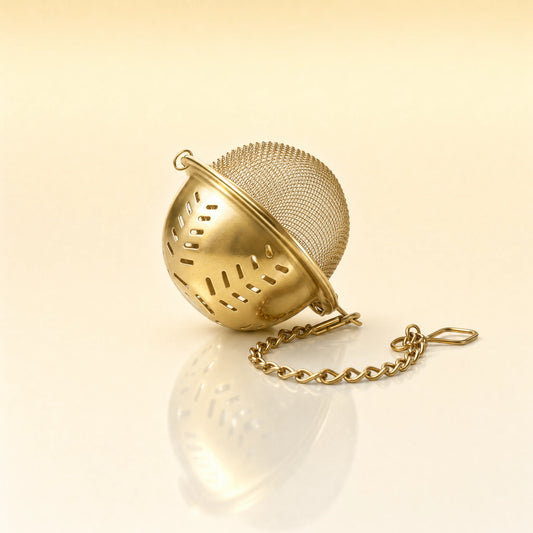 GOLDEN LEAF STRAINER - A gold-finished metal tea infuser ball with a matching chain, resting on a reflective cream surface.