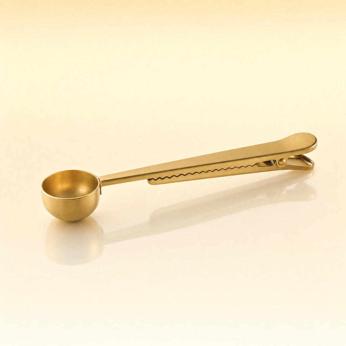 Golden Stir Spoon – Gold-toned stainless steel tea scoop with a round bowl and a long flat handle with a built-in clip, photographed on a warm cream background with a soft reflection below.