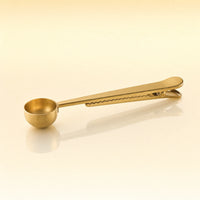 Golden Stir Spoon – Gold-toned stainless steel tea scoop with a round bowl and a long flat handle with a built-in clip, photographed on a warm cream background with a soft reflection below.