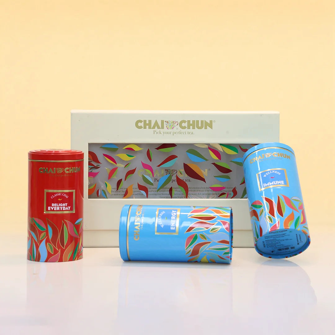 A set of ChaiChun tea tins and a gift box on a white reflective surface against a pale yellow background. The set includes a red tin labeled 'Delicious Everyday,' a blue tin labeled 'Magazine,' a blue cylindrical tin, and a white box with a colorful leaf pattern and the ChaiChun brand name.