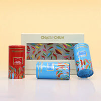 A set of ChaiChun tea tins and a gift box on a white reflective surface against a pale yellow background. The set includes a red tin labeled 'Delicious Everyday,' a blue tin labeled 'Magazine,' a blue cylindrical tin, and a white box with a colorful leaf pattern and the ChaiChun brand name.