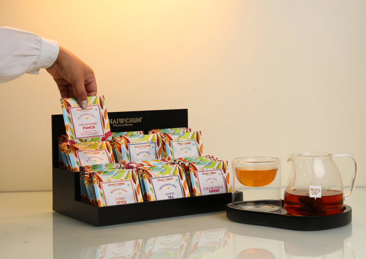 Guest-Room - A hand wearing a white sleeve reaches into a black display box filled with colorful tea bags from the brand Hairichun. To the right, a glass cup and a glass teapot with amber tea sit on a black tray against a warm beige background.