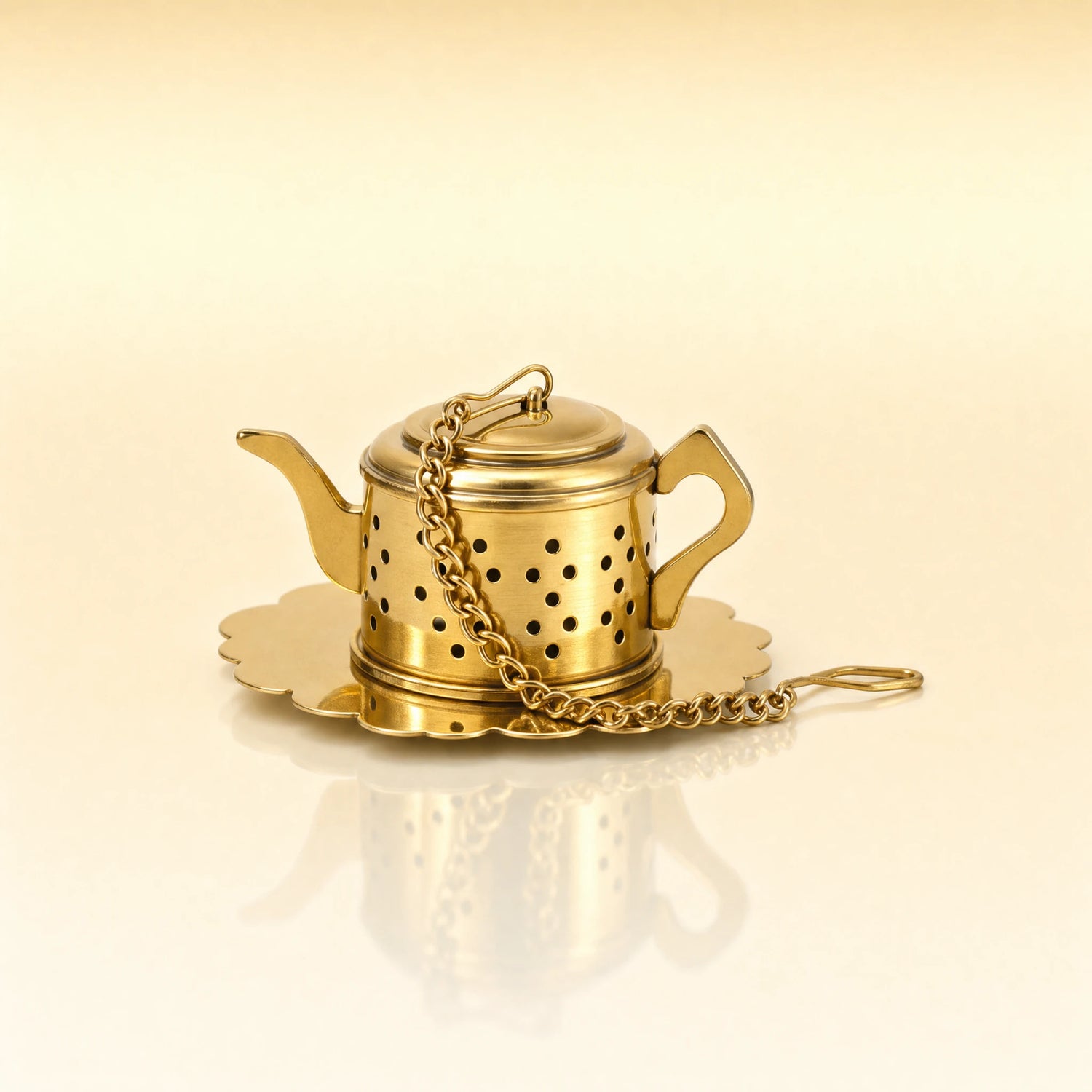 IMPERIAL KETTLE INFUSER - A miniature gold-toned metal teapot-shaped tea infuser with a matching scalloped saucer and chain, photographed on a warm cream background with a soft reflection below.