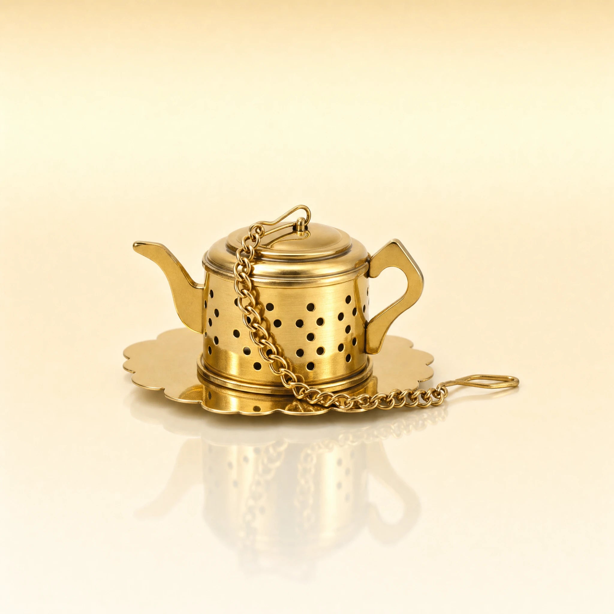 IMPERIAL KETTLE INFUSER - A miniature gold-toned metal teapot-shaped tea infuser with a matching scalloped saucer and chain, photographed on a warm cream background with a soft reflection below.