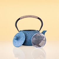 IceVineIronTeapot2 - A blue cast iron teapot with a crackle glaze, featuring a black handle wrapped with beige cord, shown with its lid and glass cover on a reflective surface against a warm beige background.