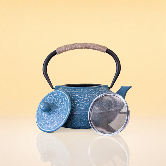 IceVineIronTeapot2 - A blue cast iron teapot with a crackle glaze, featuring a black handle wrapped with beige cord, shown with its lid and glass cover on a reflective surface against a warm beige background.