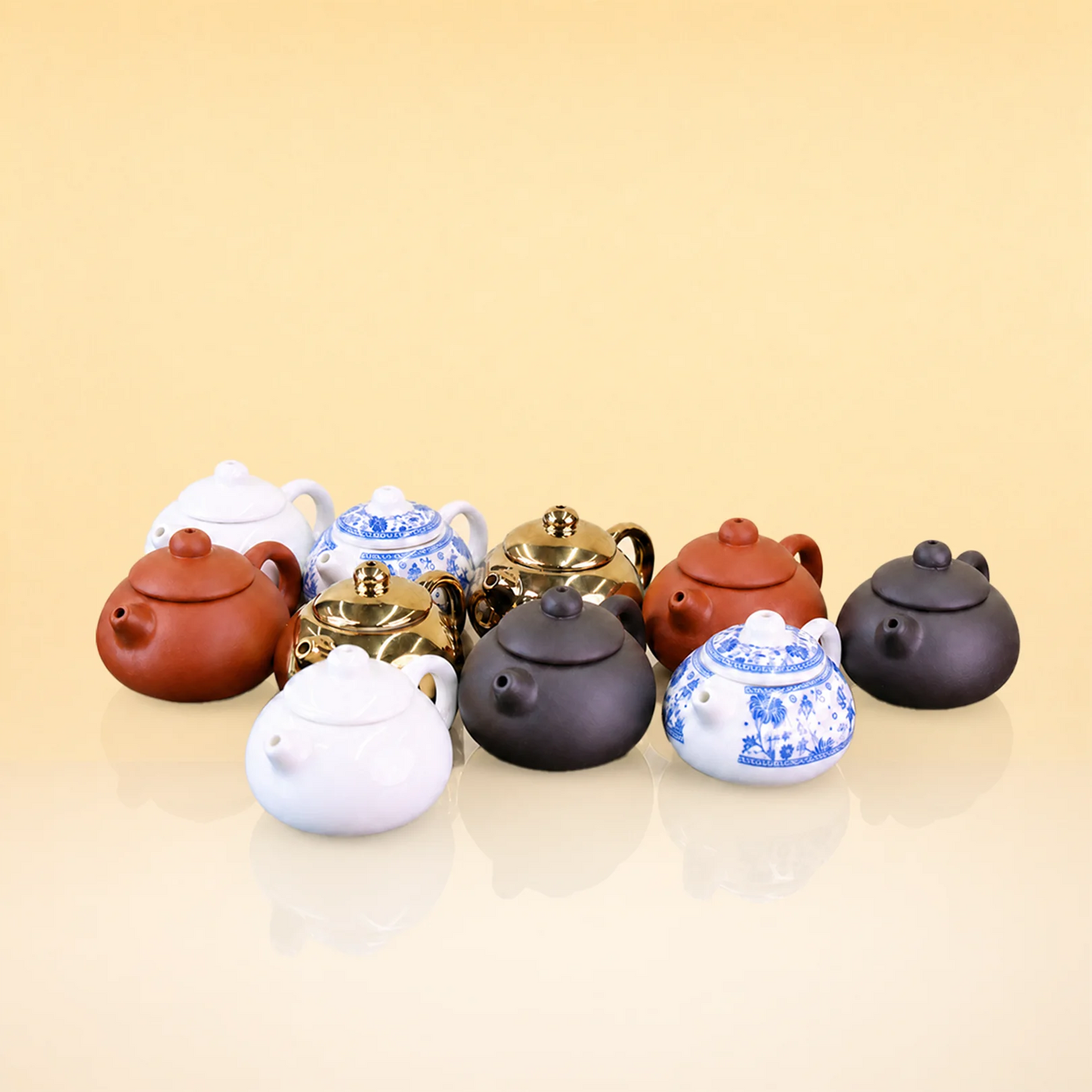 ImperialPot - A group of miniature ceramic teapots in various colors and patterns, including solid brown, white, black, gold, and blue-and-white floral designs, arranged on a reflective beige surface against a solid beige background.