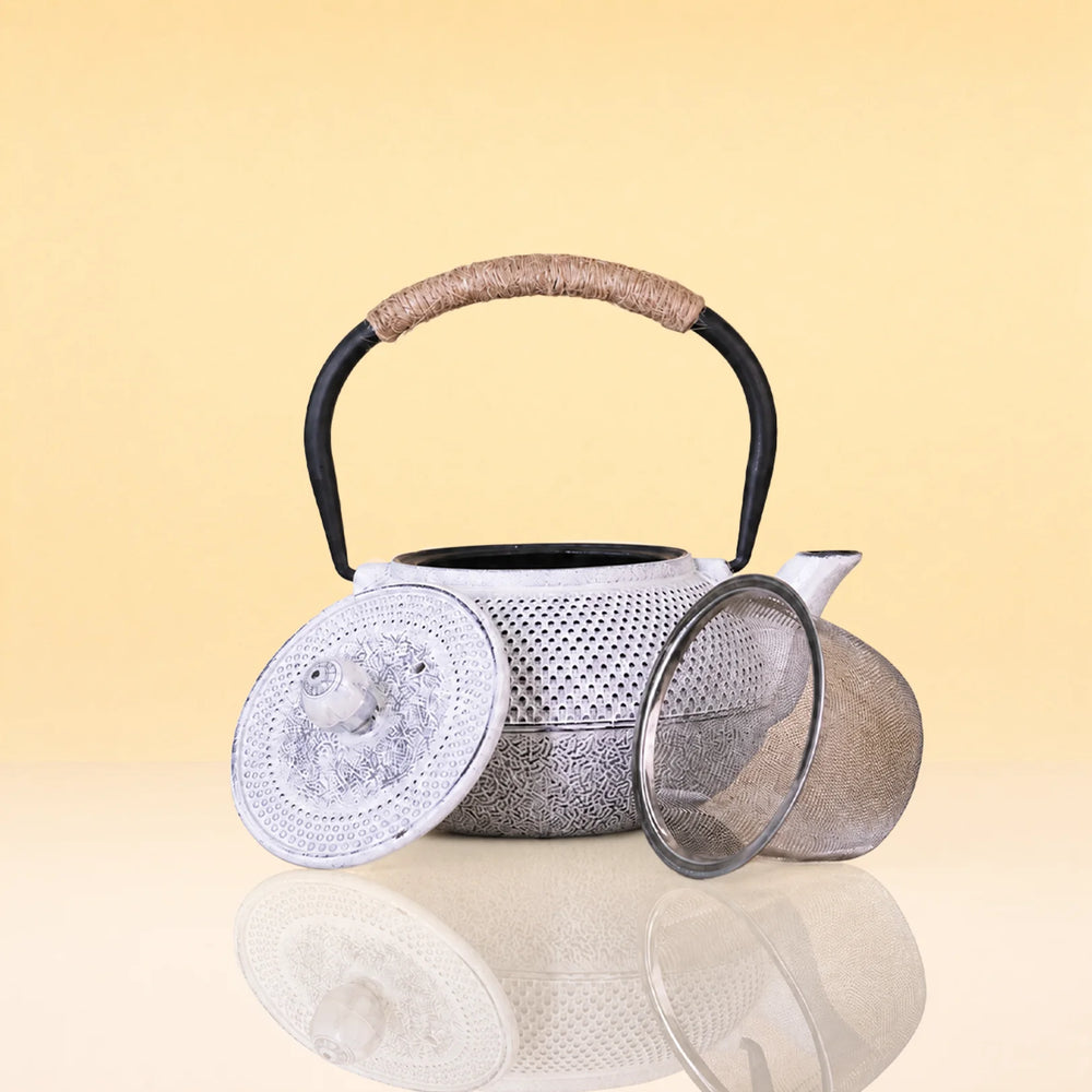 ImpreialMistIronTeapot - A silver metal teapot with a black handle and a wrapped beige handle grip, shown with its lid and mesh strainer on a reflective surface against a warm yellow background.