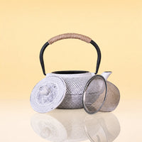 ImpreialMistIronTeapot - A silver metal teapot with a black handle and a wrapped beige handle grip, shown with its lid and mesh strainer on a reflective surface against a warm yellow background.