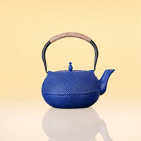 IndigoFlushIronTeapot - A blue cast iron teapot with a textured surface, a black metal handle wrapped with beige rope, and a beige spout, shown on a reflective white surface against a warm beige background.