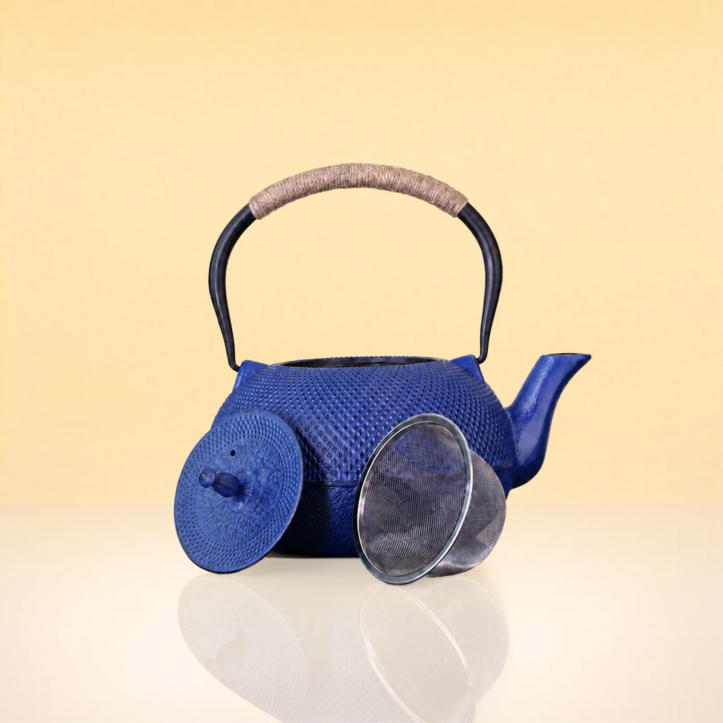 IndigoFlushIronTeapot2 - A blue cast iron teapot with a textured woven pattern, a dark metal handle wrapped in beige fabric, and a matching blue lid leaning against the front, set on a reflective white surface against a warm beige background.