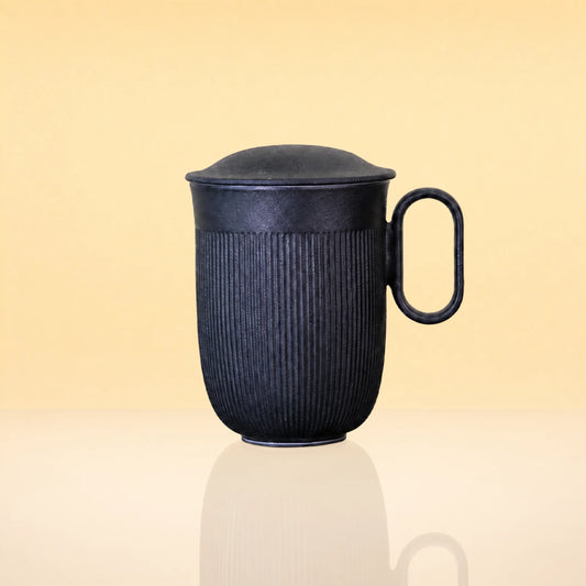 InfusionMug-CeramicTeaMugwithanInfuserandLid - A dark charcoal gray ribbed ceramic mug with a matching lid and a loop handle, shown on a reflective cream surface against a warm beige background.