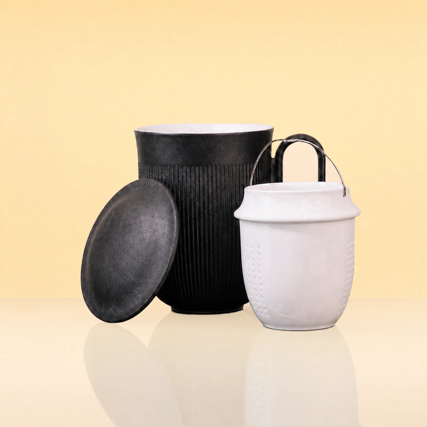 InfusionMug-CeramicTeaMugwithanInfuserandLid2 - Two ceramic canisters with lids on a warm beige background. One canister is black with a ribbed texture and a separate matching lid leaning against it.