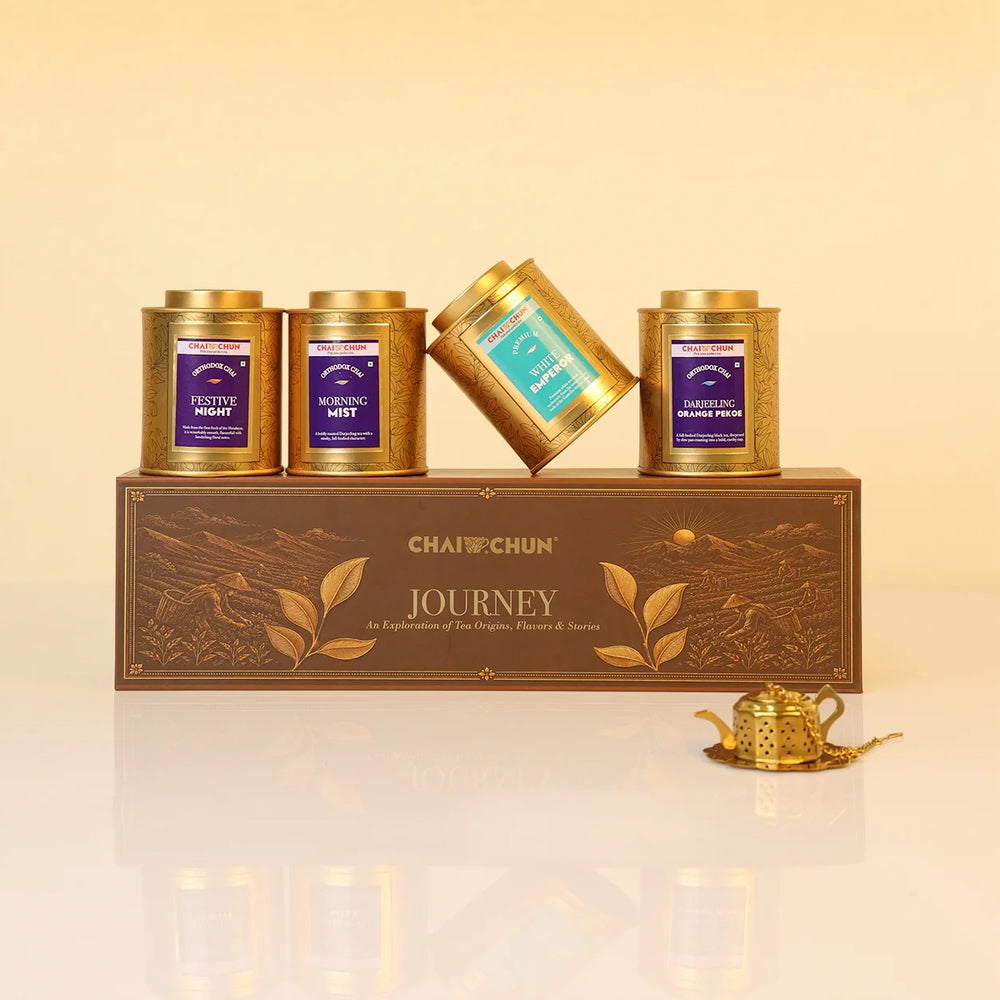 Journey-Collection-v2 - A boxed tea gift set from CHAI CHUNJUN named JOURNEY, featuring four gold metal tea tins with purple and teal labels arranged on and around a brown decorative box, with a small gold teapot on a reflective white surface against a warm beige background.