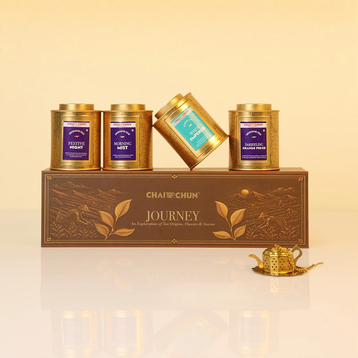 Journey-Collection-v2 - A boxed tea gift set from CHAI CHUNJUN named JOURNEY, featuring four gold metal tea tins with purple and teal labels arranged on and around a brown decorative box, with a small gold teapot on a reflective white surface against a warm beige background.