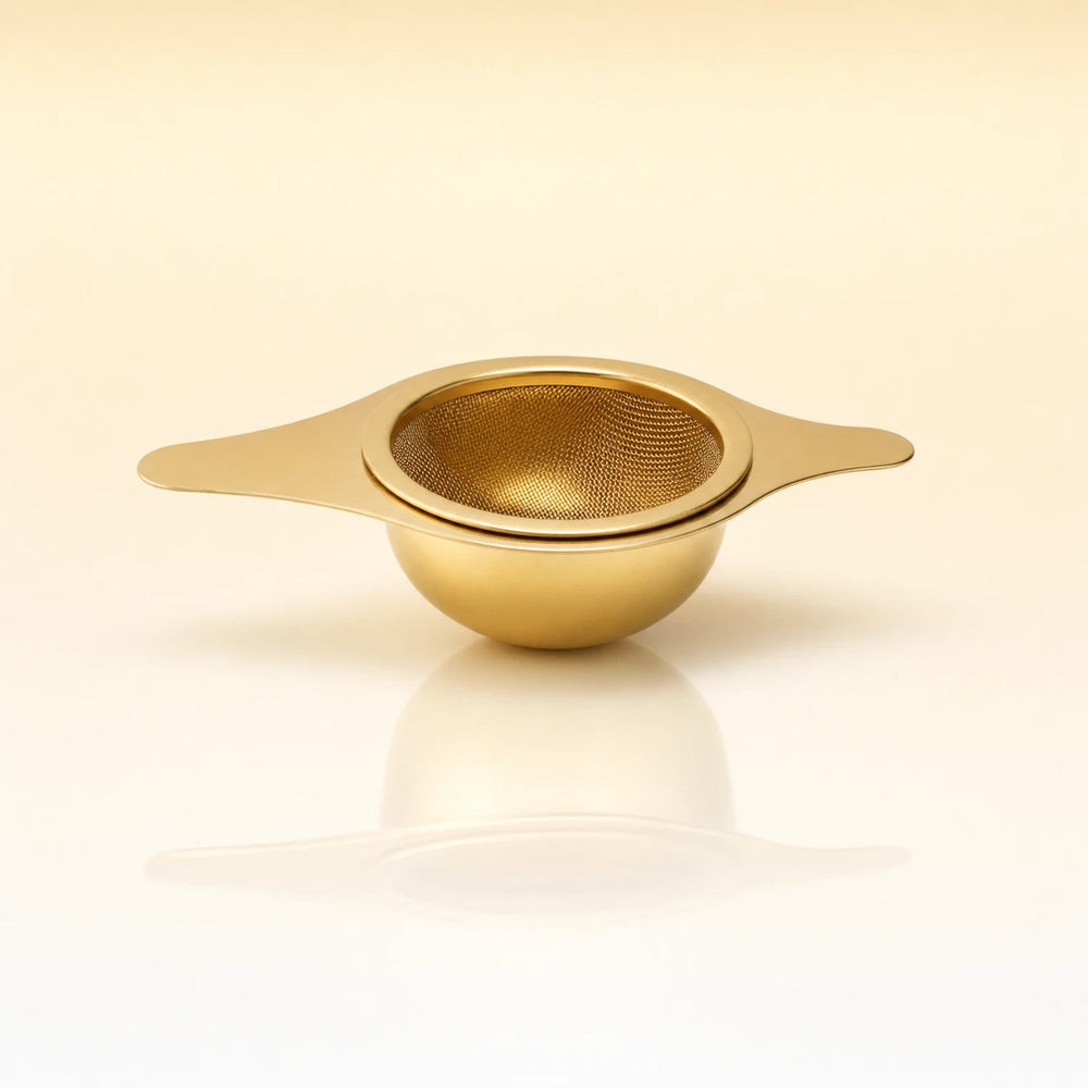 Lotus Bowl Infuser – Gold-toned stainless steel shallow bowl-shaped tea infuser with two flat side handles, photographed on a warm cream background with a soft reflection below.