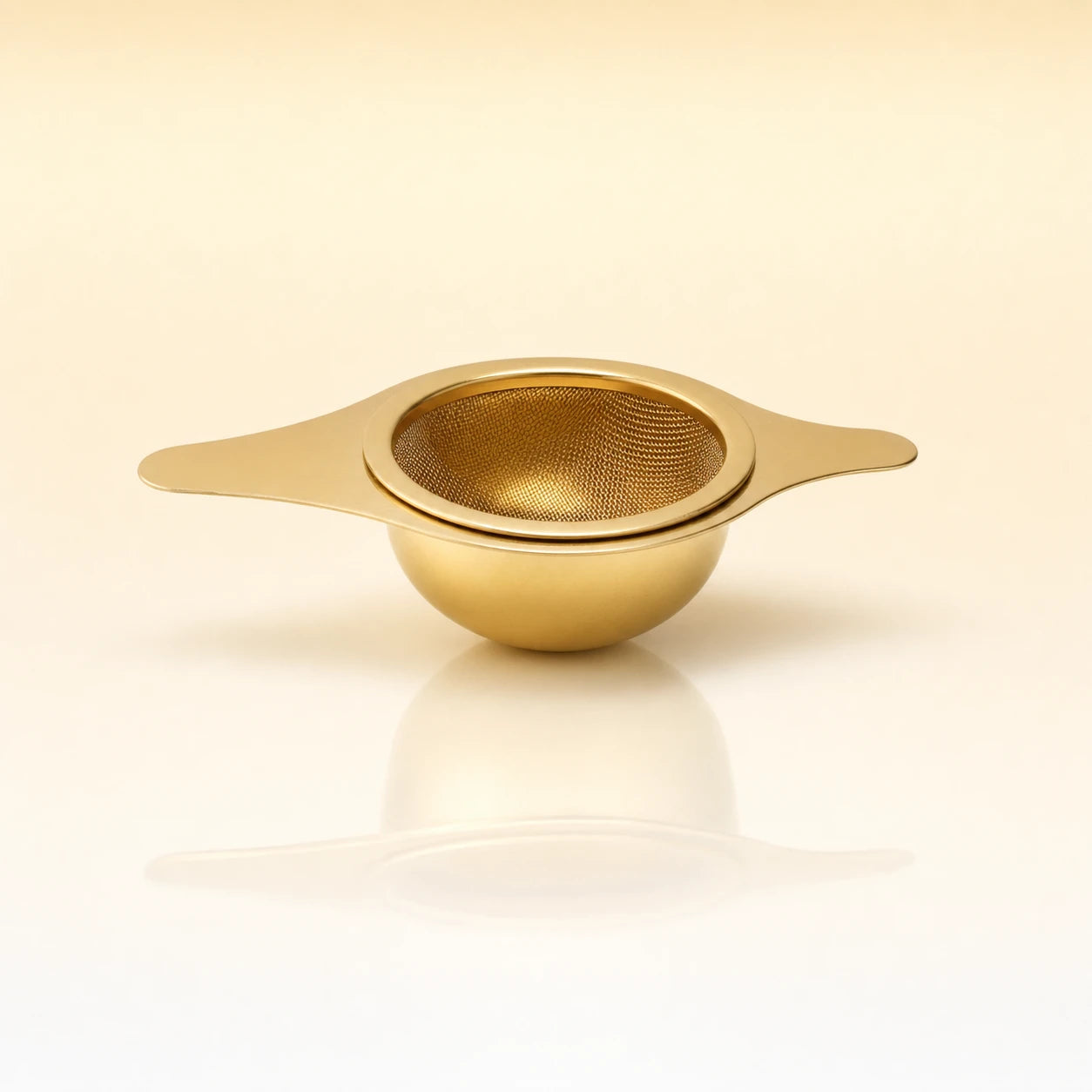 Lotus Bowl Infuser – Gold-toned stainless steel shallow bowl-shaped tea infuser with two flat side handles, photographed on a warm cream background with a soft reflection below.