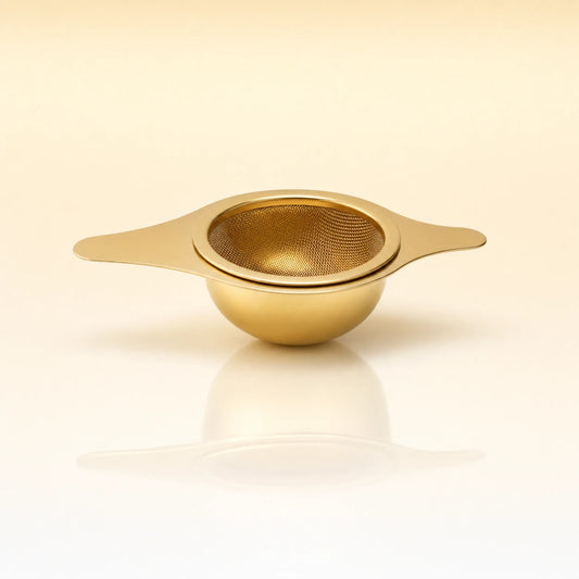 Lotus Bowl Infuser – Gold-toned stainless steel shallow bowl-shaped tea infuser with two flat side handles, photographed on a warm cream background with a soft reflection below.