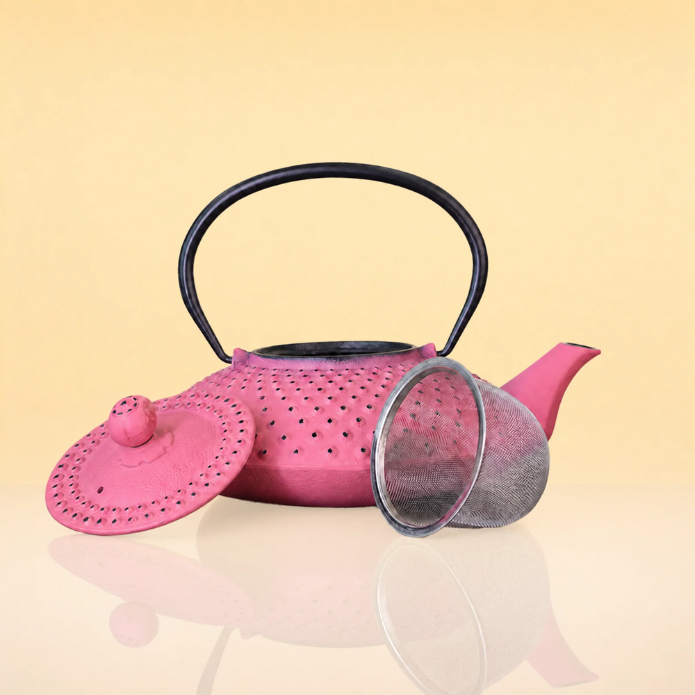 MagentaSteepIronTeapot2 - A pink perforated metal teapot with a black handle, shown with its matching pink lid and a silver mesh strainer on a reflective beige surface against a pale yellow background.