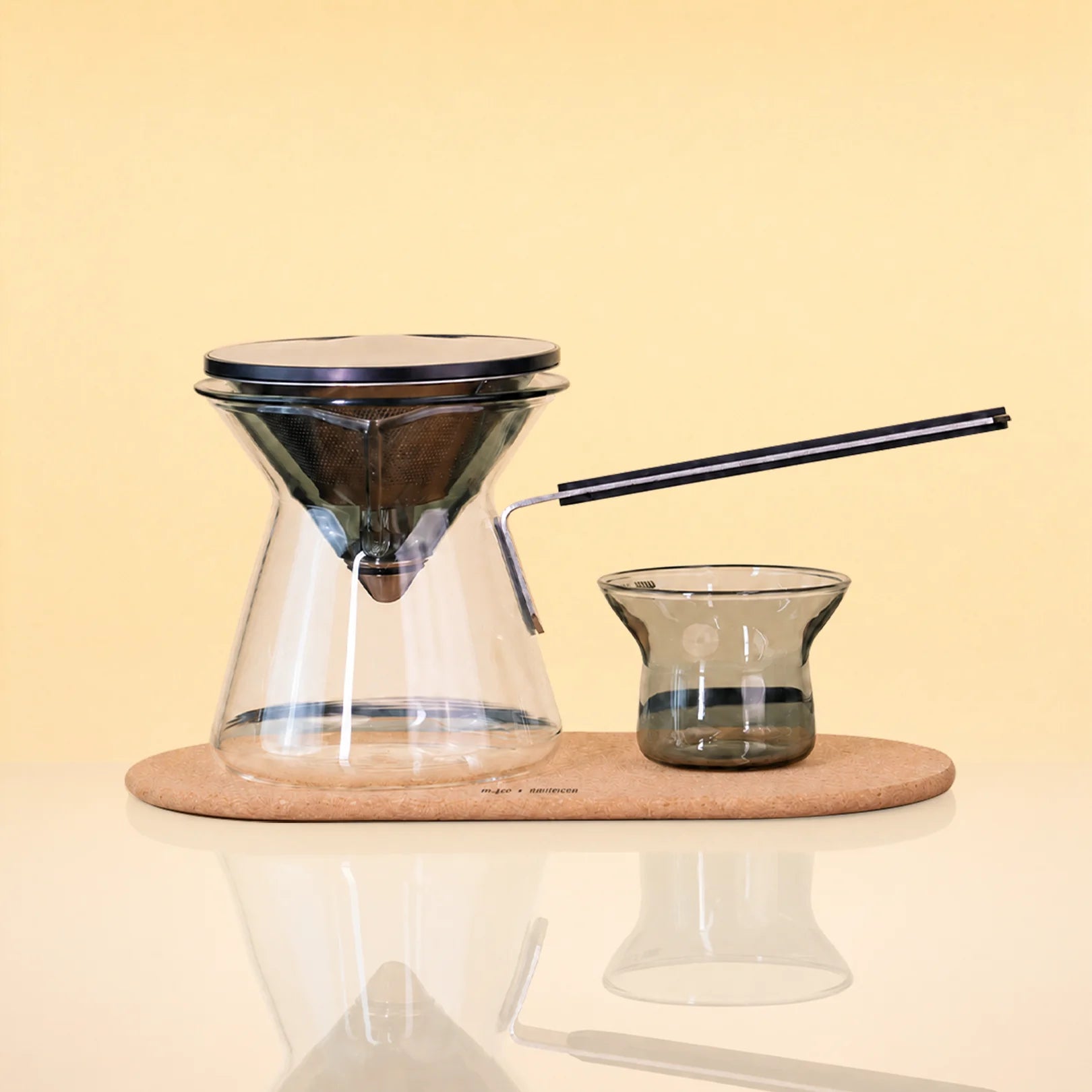 ObsidianEditionCeremonySet - A pour-over coffee set on a light wood tray, featuring a clear glass cone-shaped dripper with a black rim, a matching glass carafe, a small dark glass cup, and a black metal straw, photographed against a warm beige background.