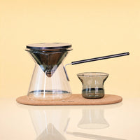 ObsidianEditionCeremonySet - A pour-over coffee set on a light wood tray, featuring a clear glass cone-shaped dripper with a black rim, a matching glass carafe, a small dark glass cup, and a black metal straw, photographed against a warm beige background.