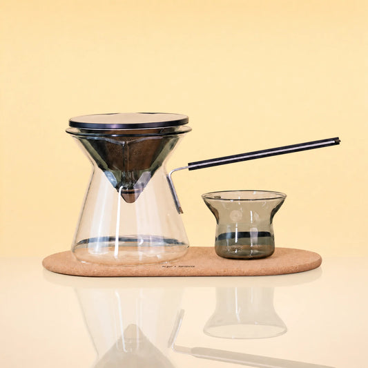 ObsidianEditionCeremonySet - A pour-over coffee set on a light wood tray, featuring a clear glass cone-shaped dripper with a black rim, a matching glass carafe, a small dark glass cup, and a black metal straw, photographed against a warm beige background.
