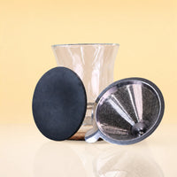 ObsidianEditionCeremonySet3 - A clear glass vase with a flared rim, shown with two accessories: a round black textured disc and a metal mesh funnel with a black rim, set against a warm beige background.