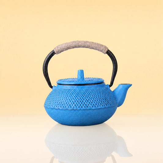 OceanicFlushTeapot - A bright blue cast iron teapot with a textured surface, a black metal handle wrapped in a beige fabric, and a matching blue lid, shown on a reflective white surface against a warm beige background.