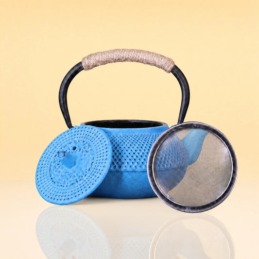 OceanicFlushTeapot2 - A blue perforated metal teapot with a black handle wrapped in beige cord, shown with its lid off and a round mesh strainer resting beside it on a reflective surface against a pale yellow background.