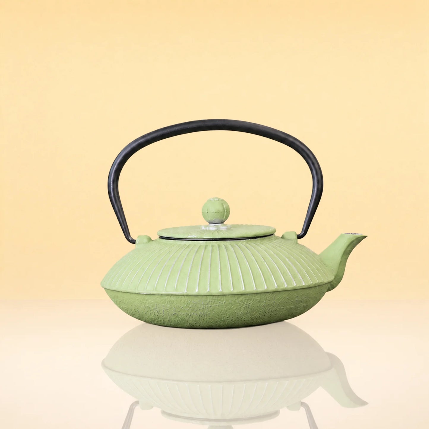 OliveInfusionTeaKettle - A light green cast iron teapot with a ribbed leaf-like pattern, a black metal handle, and a matching green lid knob, set against a warm beige gradient background with a soft reflection below.
