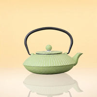 OliveInfusionTeaKettle - A light green cast iron teapot with a ribbed leaf-like pattern, a black metal handle, and a matching green lid knob, set against a warm beige gradient background with a soft reflection below.